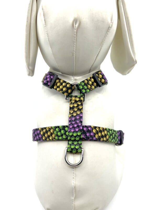 A white dog wearing a harness with purple, green and gold dots.